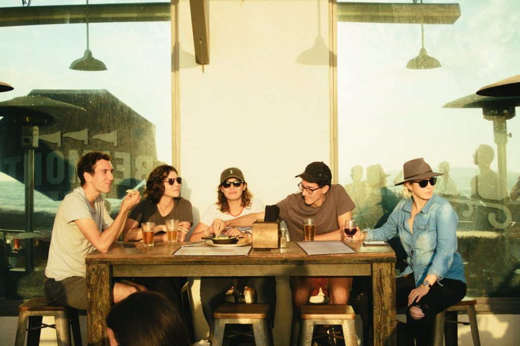 A group of five friends sits at a rustic wooden table at an outdoor restaurant during the late afternoon, enjoying drinks and food while engaged in conversation.