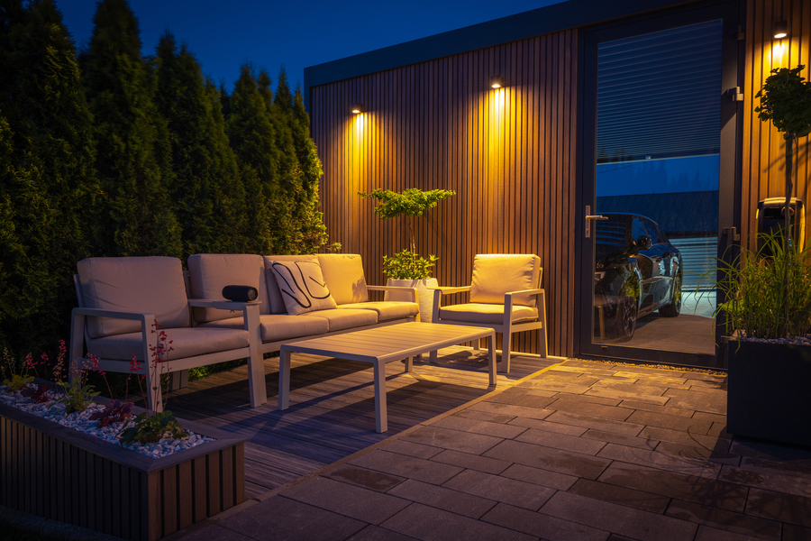 A cozy modern patio illuminated at night features cushioned seating, a minimalist table, accent lighting on wood paneling, and surrounding greenery for privacy.