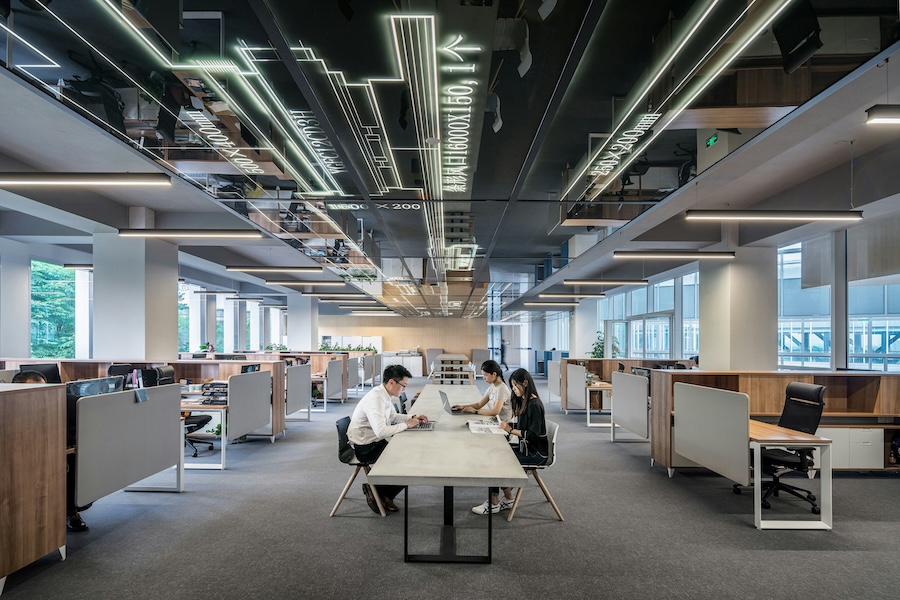 Open-plan office with workstations, partitions, and ceiling lights, featuring three colleagues collaborating at a table.
