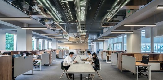 Open-plan office with workstations, partitions, and ceiling lights, featuring three colleagues collaborating at a table.