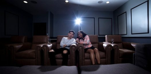 A front view of a couple shown to be sitting in a home cinema with a projector shining its light at the screen.