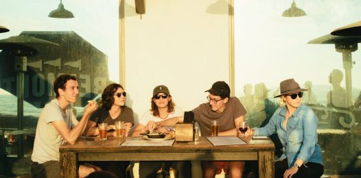 A group of five friends sits at a rustic wooden table at an outdoor restaurant during the late afternoon, enjoying drinks and food while engaged in conversation.
