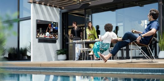 A group of people are gathered by the pool outside a house, watching a basketball game on a Samsung Terrace outdoor TV.
