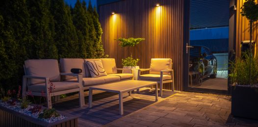A cozy modern patio illuminated at night features cushioned seating, a minimalist table, accent lighting on wood paneling, and surrounding greenery for privacy.