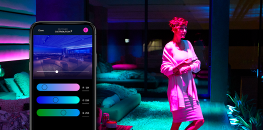 A person standing beneath blue and magenta lights. A phone is superimposed over the image, showing Savant color controls.
