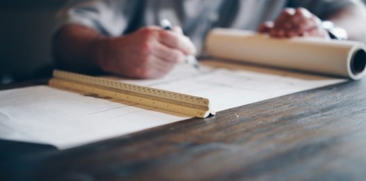 Person sitting at a table with architectural plans laid out. A ruler is in the foreground.