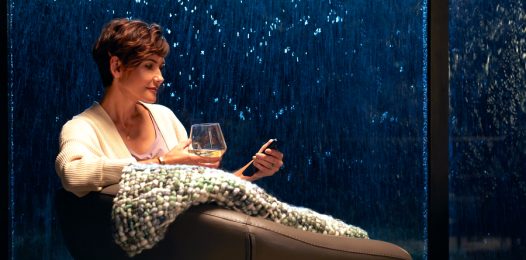 A woman sitting in a chair holding the Savant Remote with rain falling in the background.