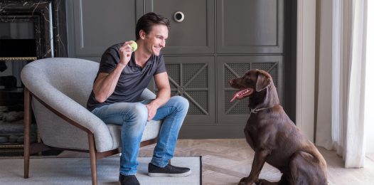 Man sitting in the living room holding a tennis ball next to his dog. On the wall is a Josh.ai microphone.