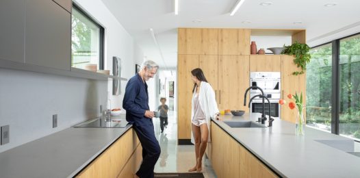 A family in a bright, modern kitchen with Savant smart home keypads on the wall.
